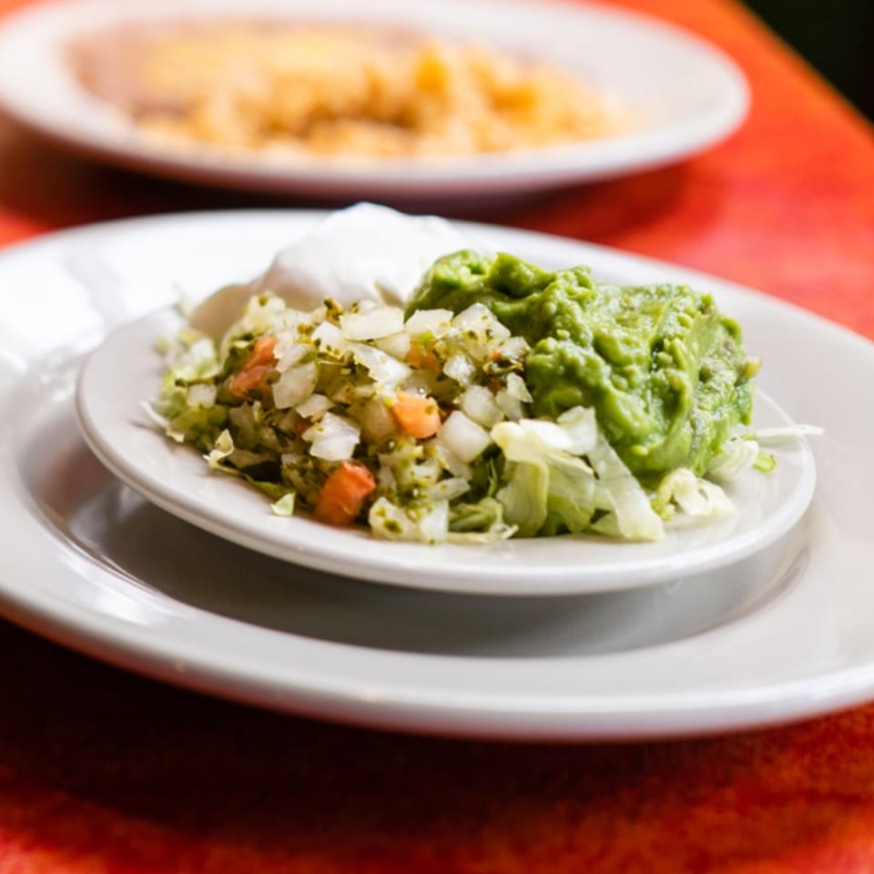 A white plate with guacamole, pico de gallo, and sour cream, with a blurred dish in the background.
